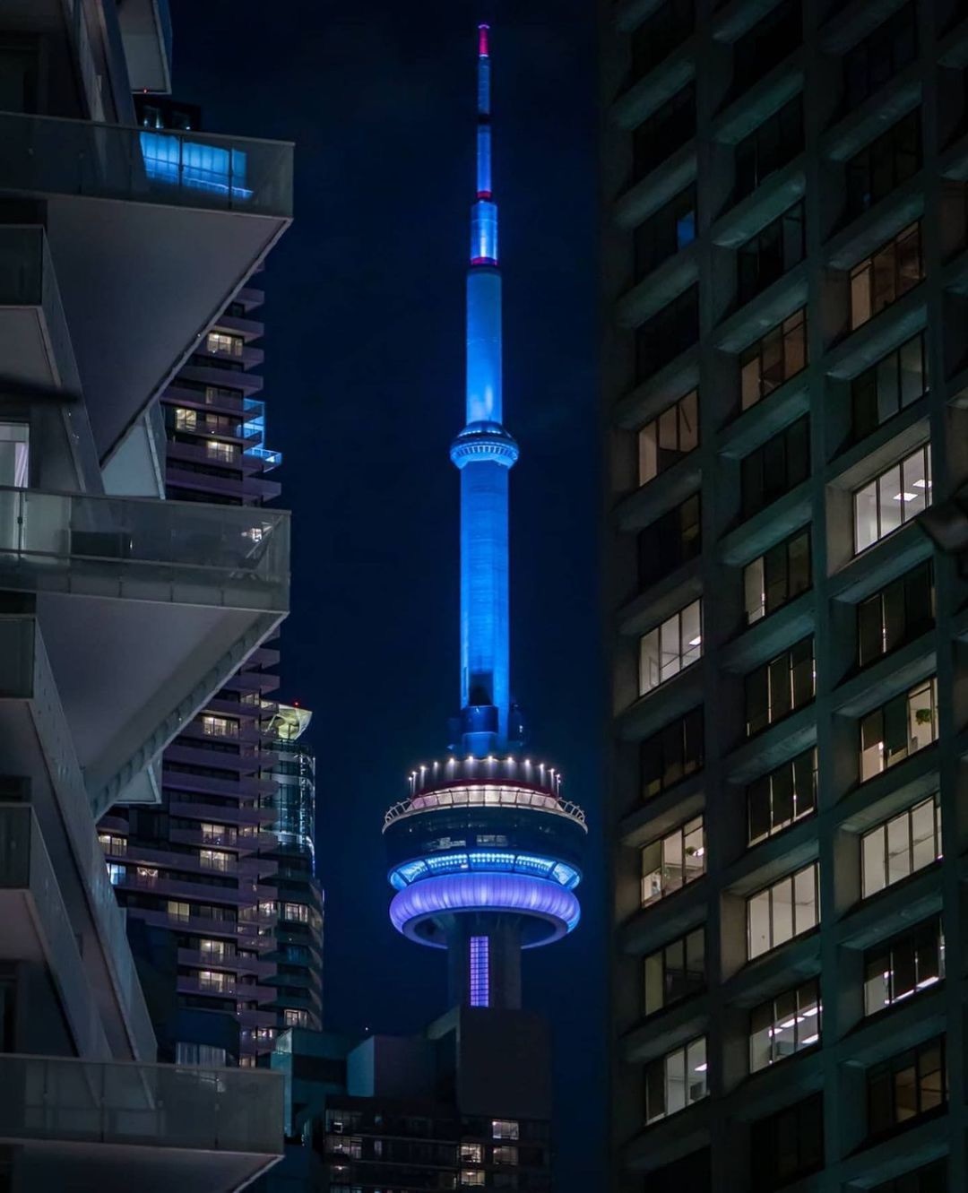 The CN Tower lit up blue between downtown towers at night